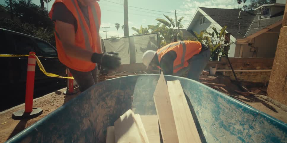 Construction workers putting pieces of wood into a small blue bucket at an outdoor construction site