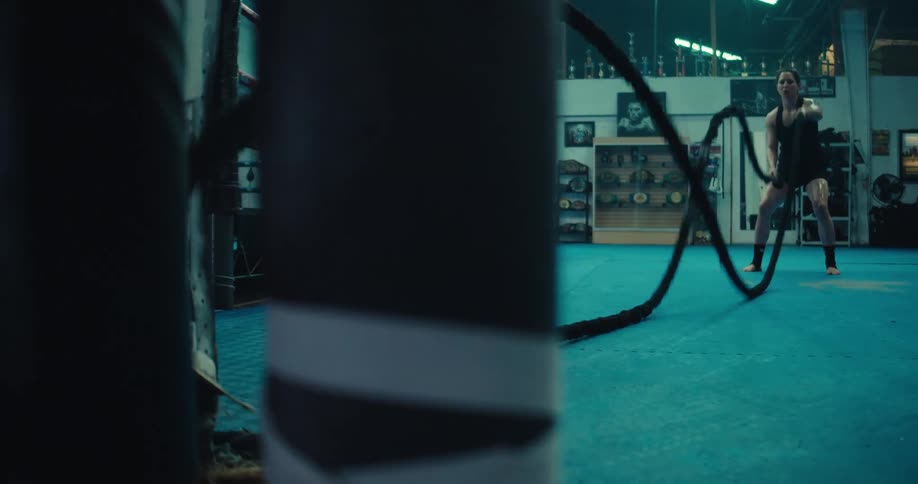 A woman is doing arm exercises with long lengths of rope in a large gym empty except for her with large fluorescent lights above her a worn blue rug under foot two punching bags and an empty boxing ring near her