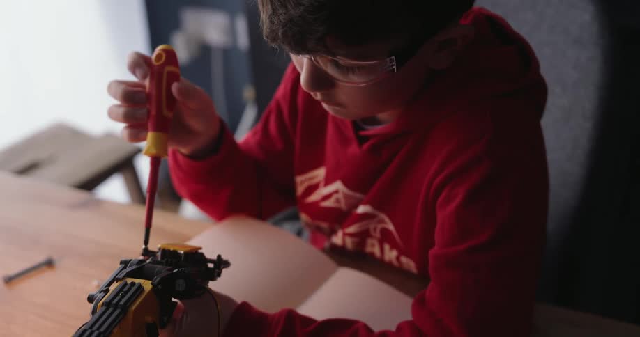 The little boy is using a screw driver on something that he is building as he sits at his desk
