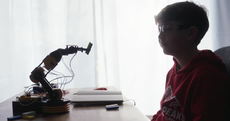 A boy in a red hoodie sits at a desk with a robotic arm wires a notebook and small electronic components illuminated by natural light