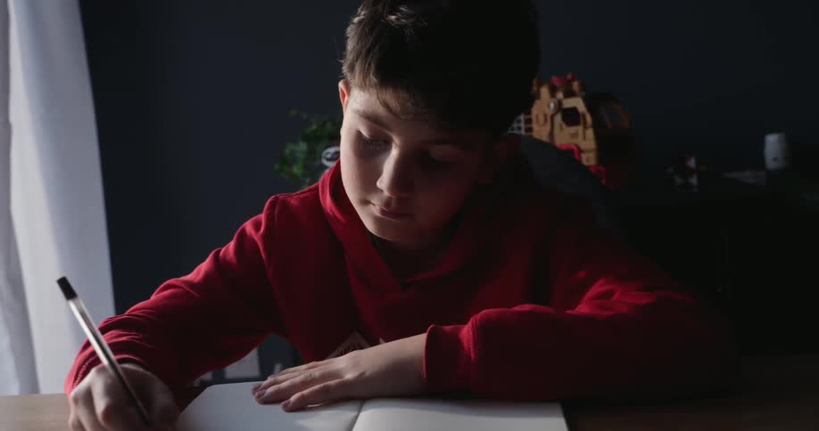 Boy sitting at a table near a curtained window writes in a notebook