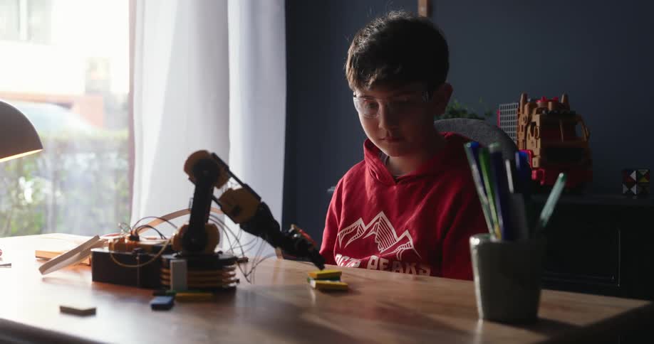 A boy in a red hoodie sits at a desk operating a robotic arm illuminated by natural light