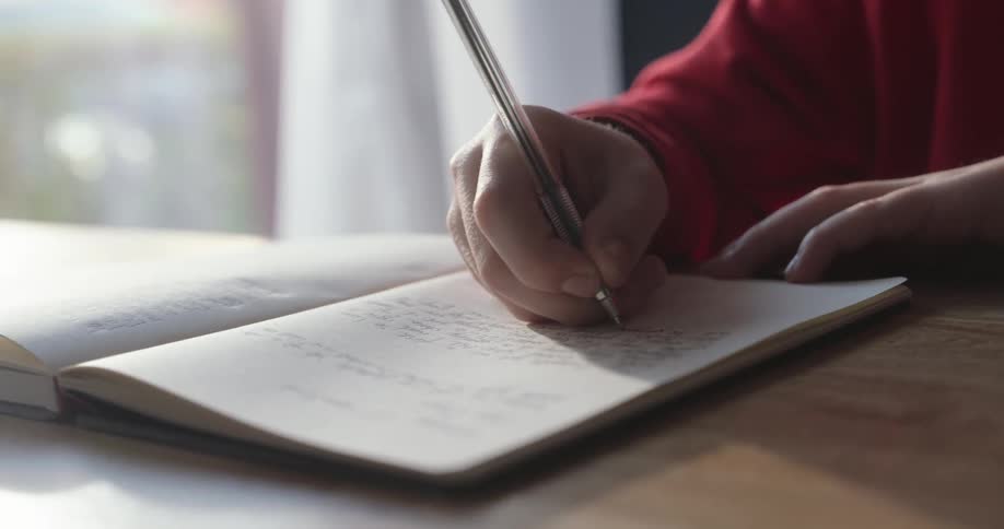 A child in a red seater is writing in a notebook with a black pen