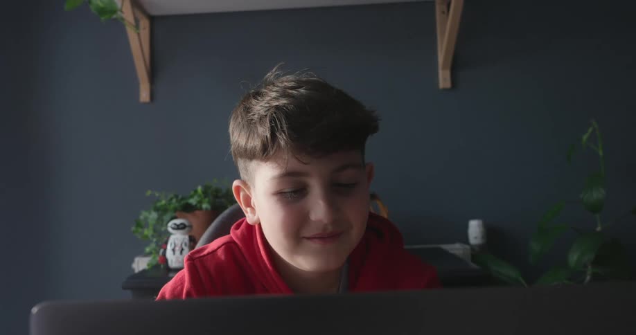A boy waves during a video call on a laptop in a room with a modern gray wall and shelves