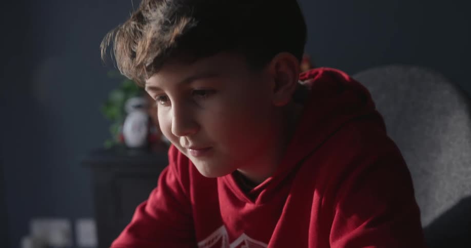 A boy in a red coat is sitting typing on a laptop and then gives a small smile