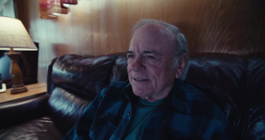 An elderly man sitting on a couch at home laughs while watching television