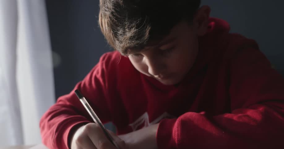 A boy in a red hoodie sits at a desk by a window writing in a notebook