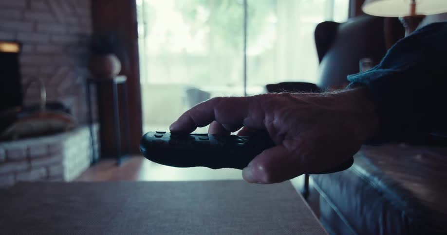 A rugged hand holding a remote control in a dimly lit living room with a blurred background featuring a fireplace a lamp and a large window letting in soft natural light