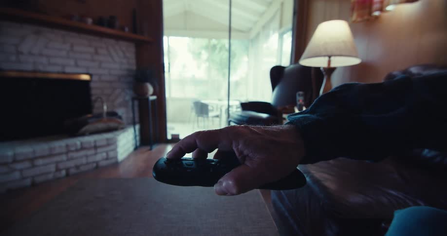 An elderly mans hand holding a remote control with a fireplace and couch visible in the background