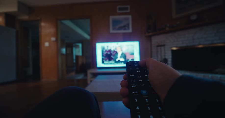A man sitting on the couch in his living room is flipping through tv channels using a remote