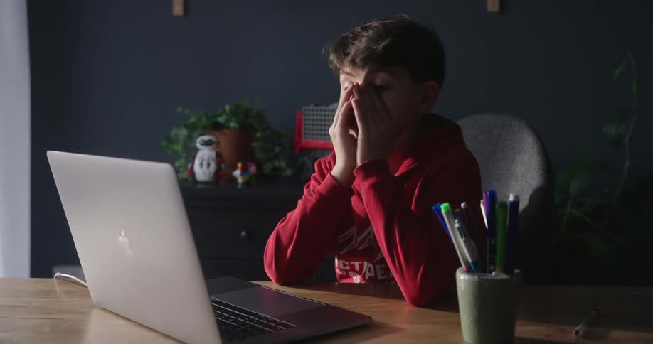 A boy wearing a red hoodie is sitting at a desk typing on his apple computer