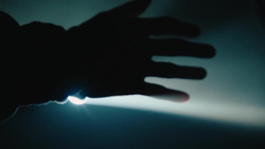 A silhouetted hand reaches into a dimly lit space its fingertips faintly illuminated by a soft glow
