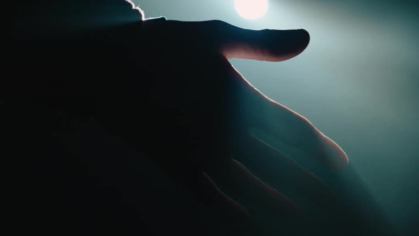 A silhouetted hand reaches into a dimly lit space its fingertips faintly illuminated by a soft glow