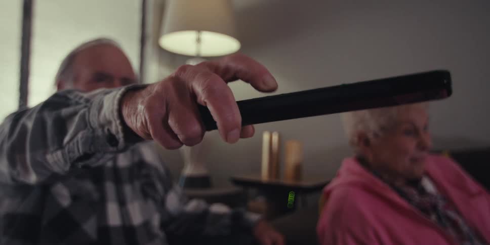 Elderly man uses the remote while he and an elderly woman sit in the living room watching television