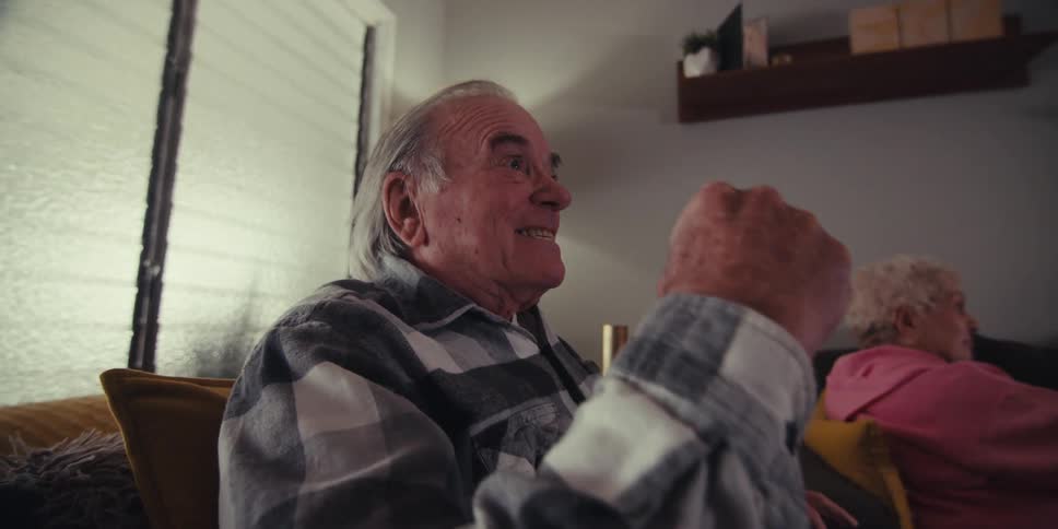 An elderly man excitedly clenches his fist while watching something with an older woman sitting beside him looking on with a more passive expression
