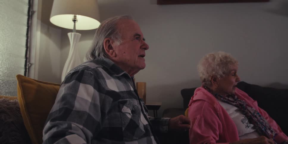 An elderly man and woman are sitting on a couch staring straight ahead and cheering and then they start to get happy