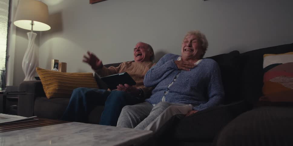Elderly couple sitting on couch laughing hysterically at something on tablet