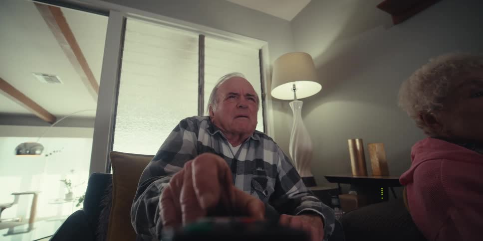 An elderly couple sits on a couch with the man picking up a remote control to change the channel both smiling and enjoying a cheerful moment