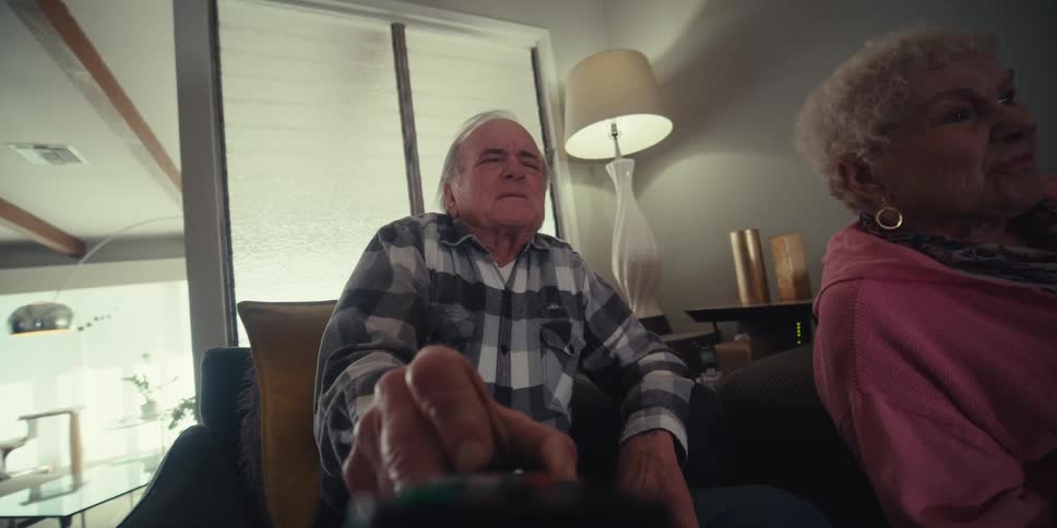 An elderly couple sits on a couch in a bright living room the man uses the tv remote control as the woman playfully takes it from his hands to use it herself