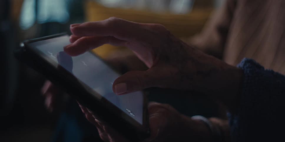 Two people sit on a couch in a dimly lit room with one gesturing toward the screen of a tablet