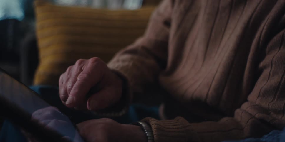 An elderly couple sits on a couch in a dimly lit room using a tablet as one of them touches the screen
