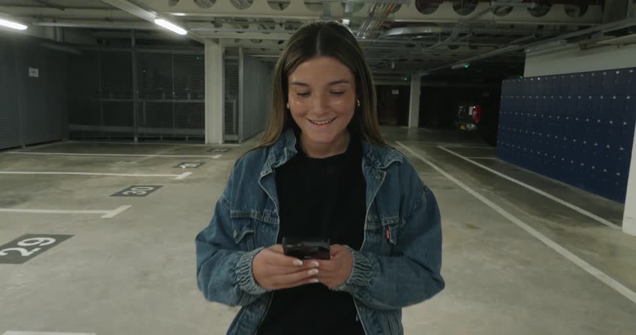 A young woman is standing with a smile on her face and is typing on her cell phone