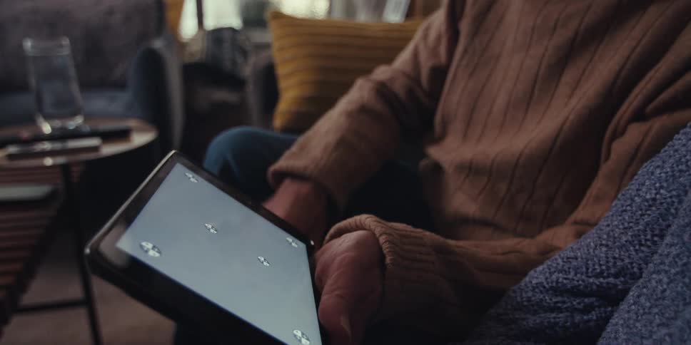 An elderly man and woman sit on a couch using a tablet and laughing in the living room