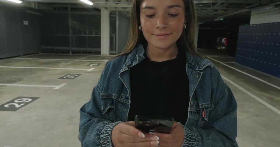 A woman wearing a denim jacket is texting on the phone while standing in an empty parking garage