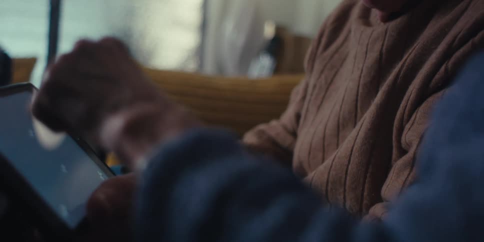 An elderly couple sits on a couch in a dimly lit room handling a tablet while conversing