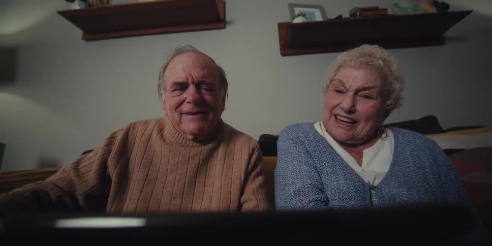An elderly couple smiles and laughs while using a tablet together in a sitting room