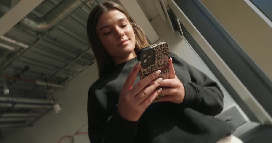 A teenage girl is standing by a window texting on her cellphone