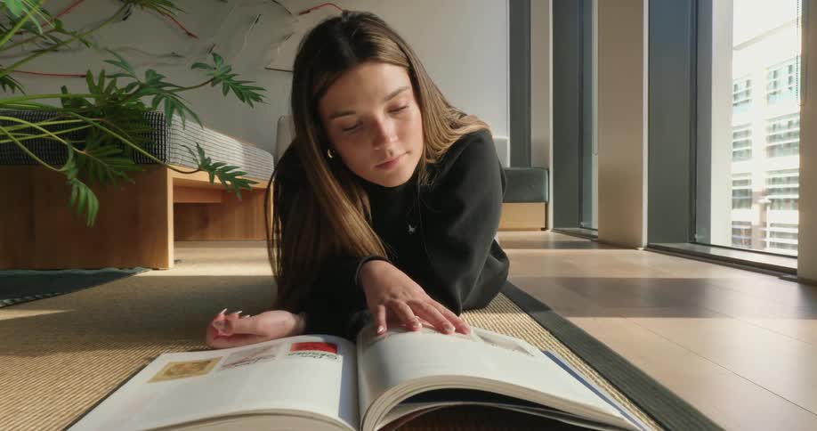 The girl is laying on the floor flipping the pages of a book