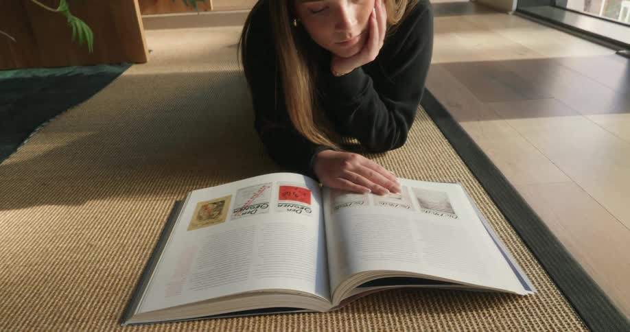 A young woman lying on a rug in a welllit room with large windows reading a large open book and resting her chin on her hand appearing focused and relaxed