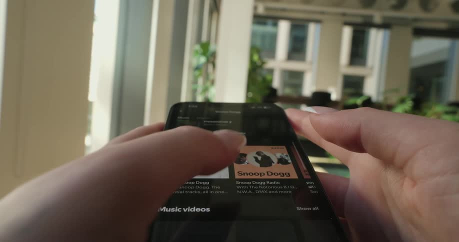 From a pov vantage a person scrolls through spotify on their phone tapping with manicured nails as they browse tracks while the bright living room in the background remains softly out of focus