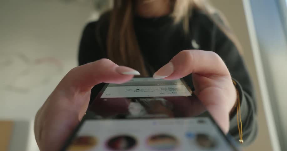 A closeup shows a persons hands holding a smartphone with their thumbs scrolling through social media