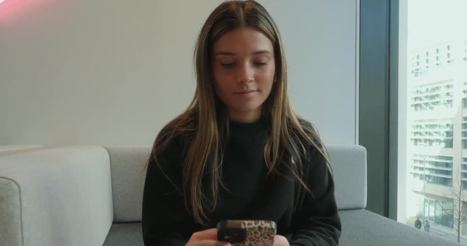 A young woman wearing a white skirt and long sleeve black shirt is using a cell phone with a stylish cover and smiling about something she sees on its screen