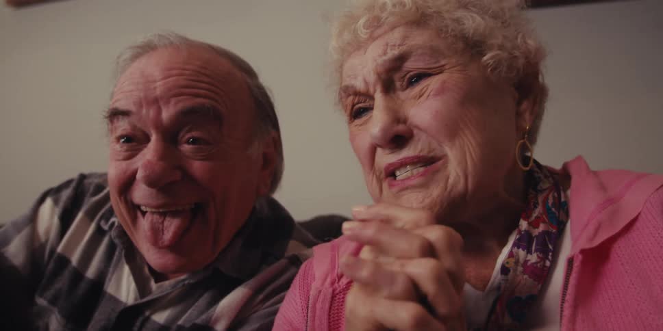 An elderly couple shares a close and playful moment with the man sticking out his tongue while the woman reacts with amusement and perhaps a touch of emotional intensity