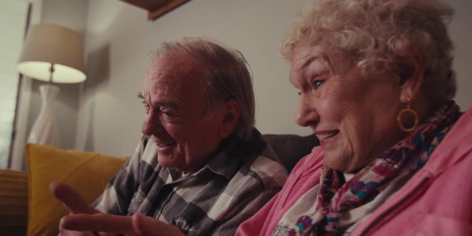 The elderly couple is sitting on the couch and the man is holding a tablet and they are both laughing and talking to it
