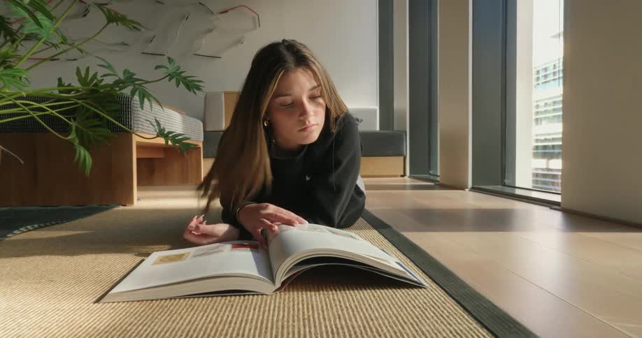 A person is lying on the floor of a sunlit room engrossed in reading an open book with a large window and a leafy plant visible in the background