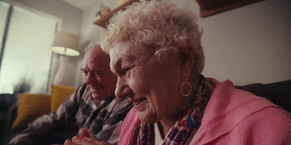 An elderly couple sits in a bright modern living room smiling and laughing as they look at a screen