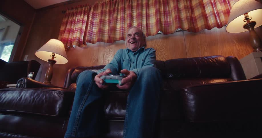 An elderly man sits on a couch at home joyfully immersed in playing video games