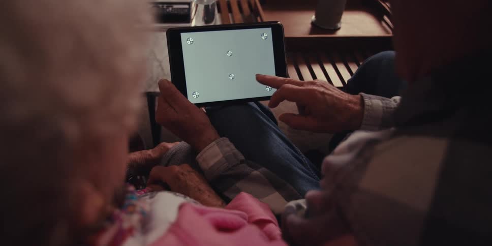 A close up shows an elderly couple seated on a couch in their living room using a tablet together