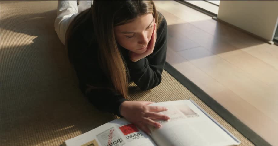 A woman reclines on a mat in a sunlit room reading an open book amidst potted plants and large windows