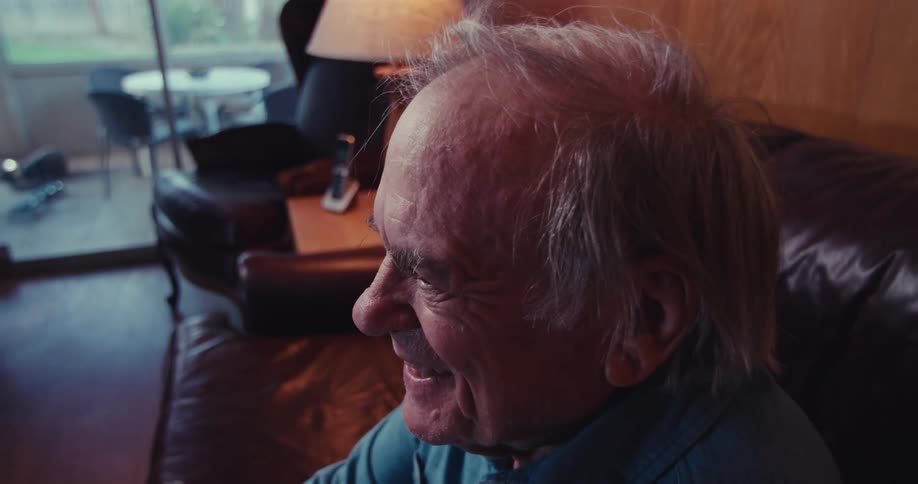An elderly man sits on a couch at home laughing and smiling while watching television
