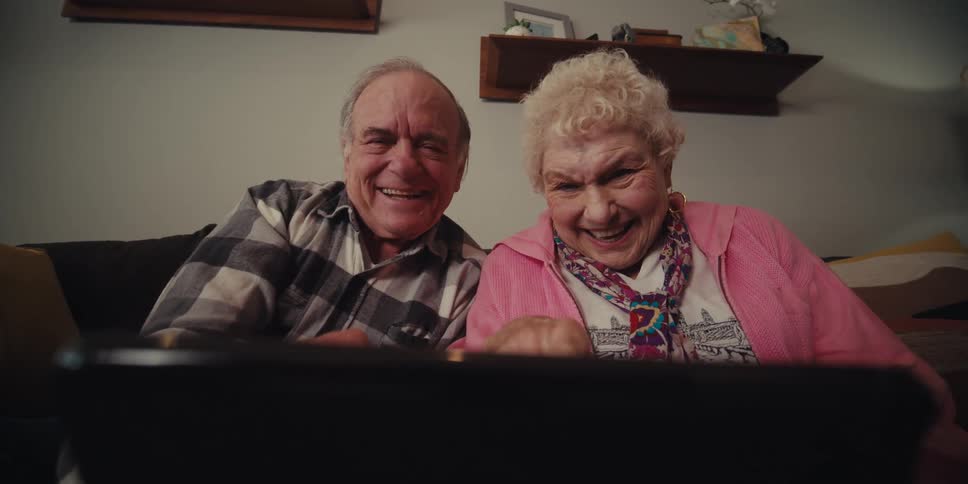 Two grandparents are talking to their grandchild on a tablet while sitting on a couch in their living room