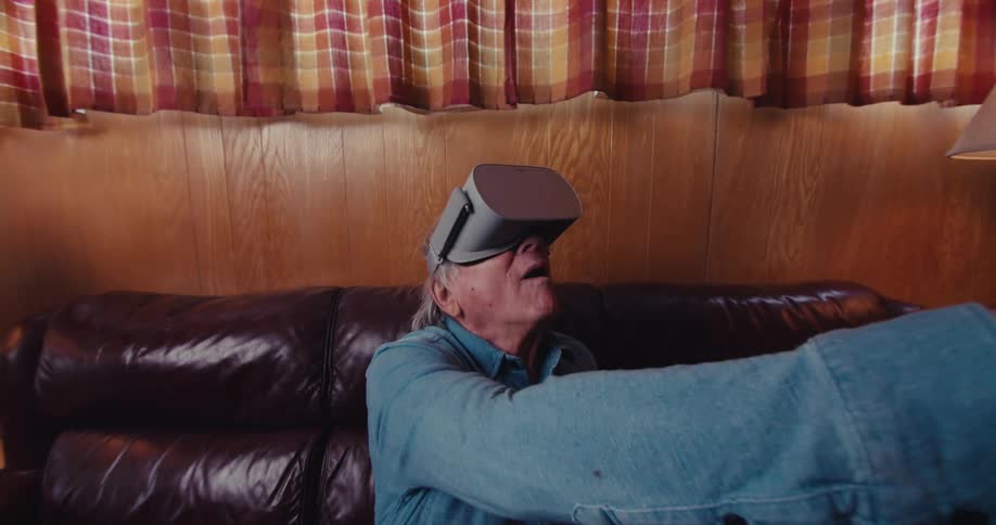 An elderly man wearing a virtual reality headset is seated on a dark brown leather couch in a cozy room with woodpaneled walls and plaid curtains