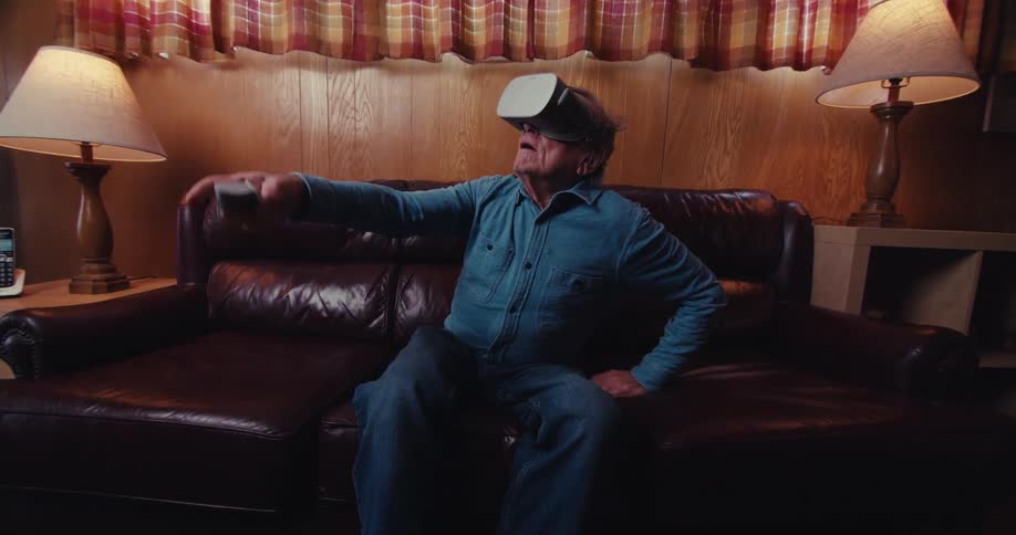An elderly man sits on a couch in the living room playing a virtual reality game while wearing goggles