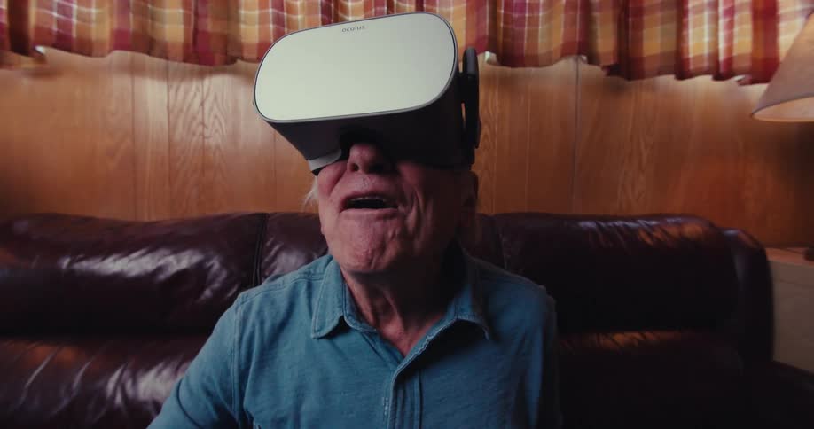 An elderly man wearing a virtual reality headset is seated on a dark brown leather couch in a cozy room with woodpaneled walls and plaid curtains
