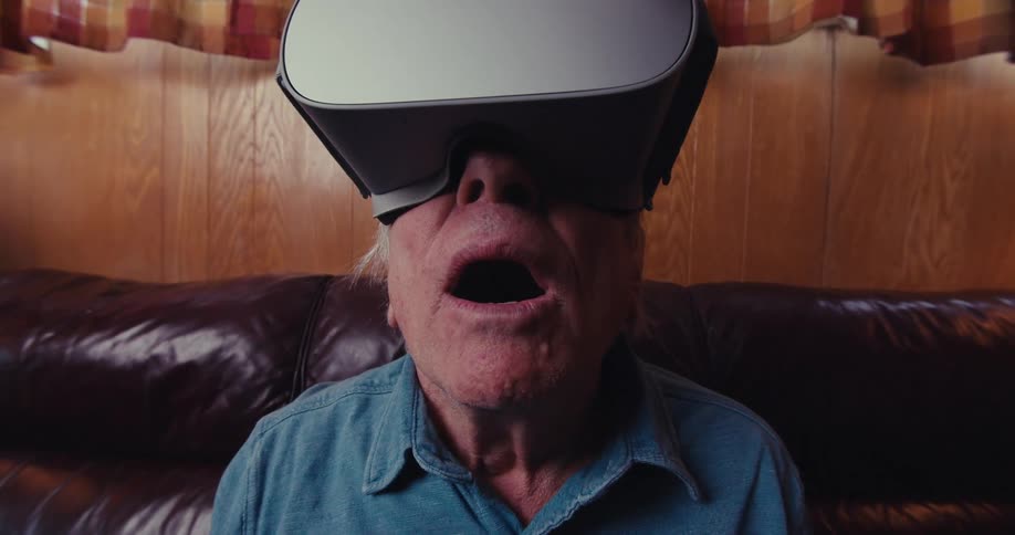 An elderly man appears captivated and surprised while experiencing virtual reality on a comfortable brown couch