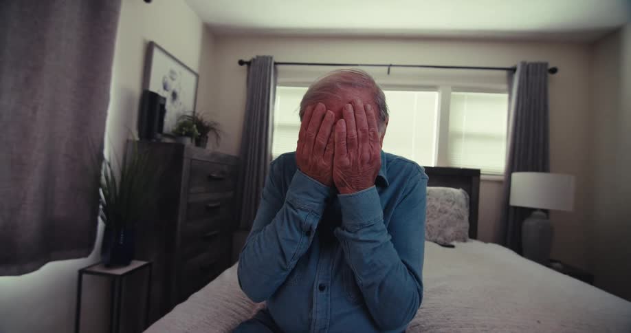 Elderly man sitting on a bed covering his face with his both his hands